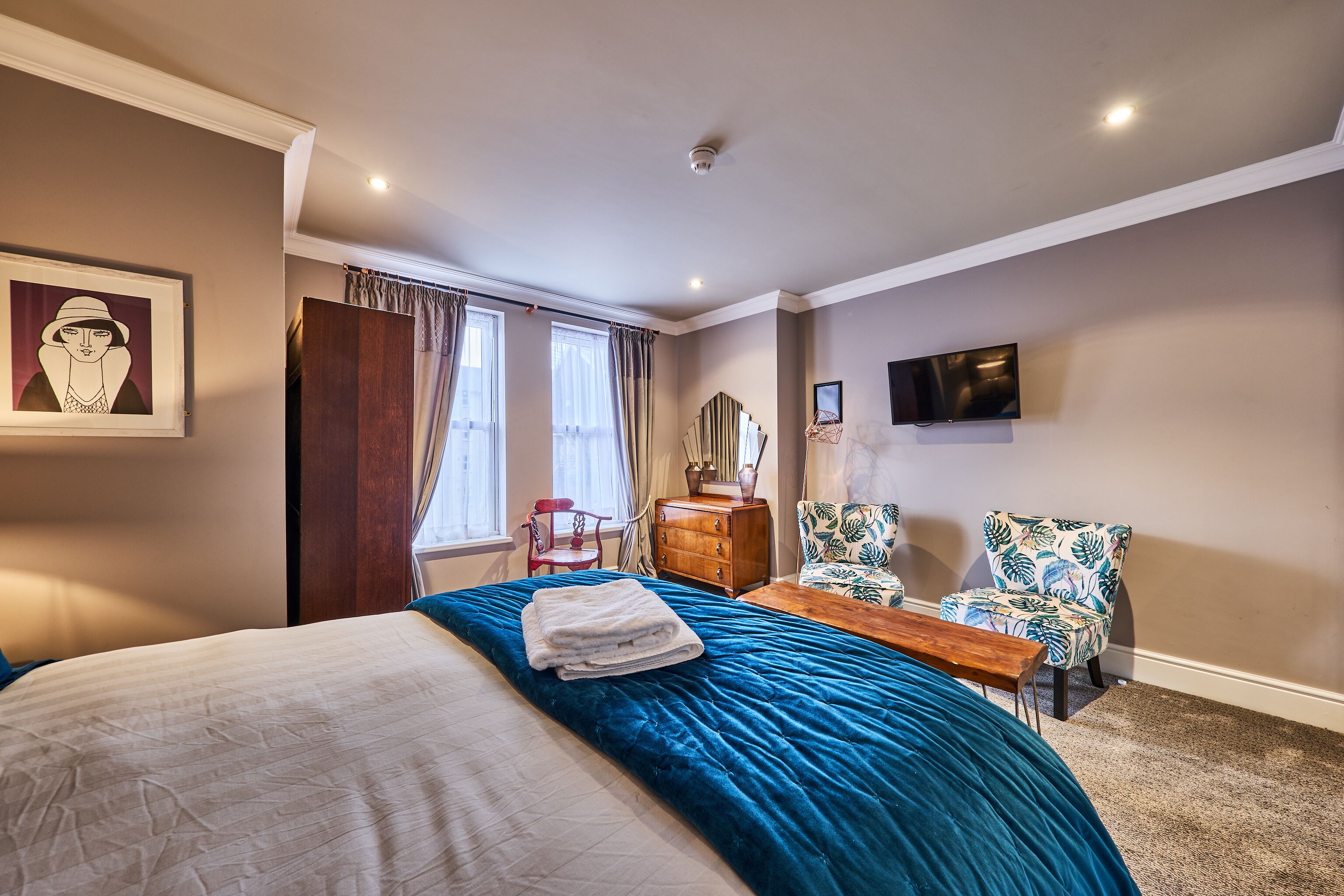 A hotel room with a large bed, a wooden dresser, a desk, a chair, a television, and a window with curtains.