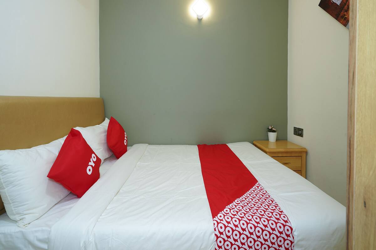 Un lit bien fait, avec une literie blanche et rouge, une tête de lit en bois, une table de chevet ornée d’une petite plante et un luminaire fixé au mur.