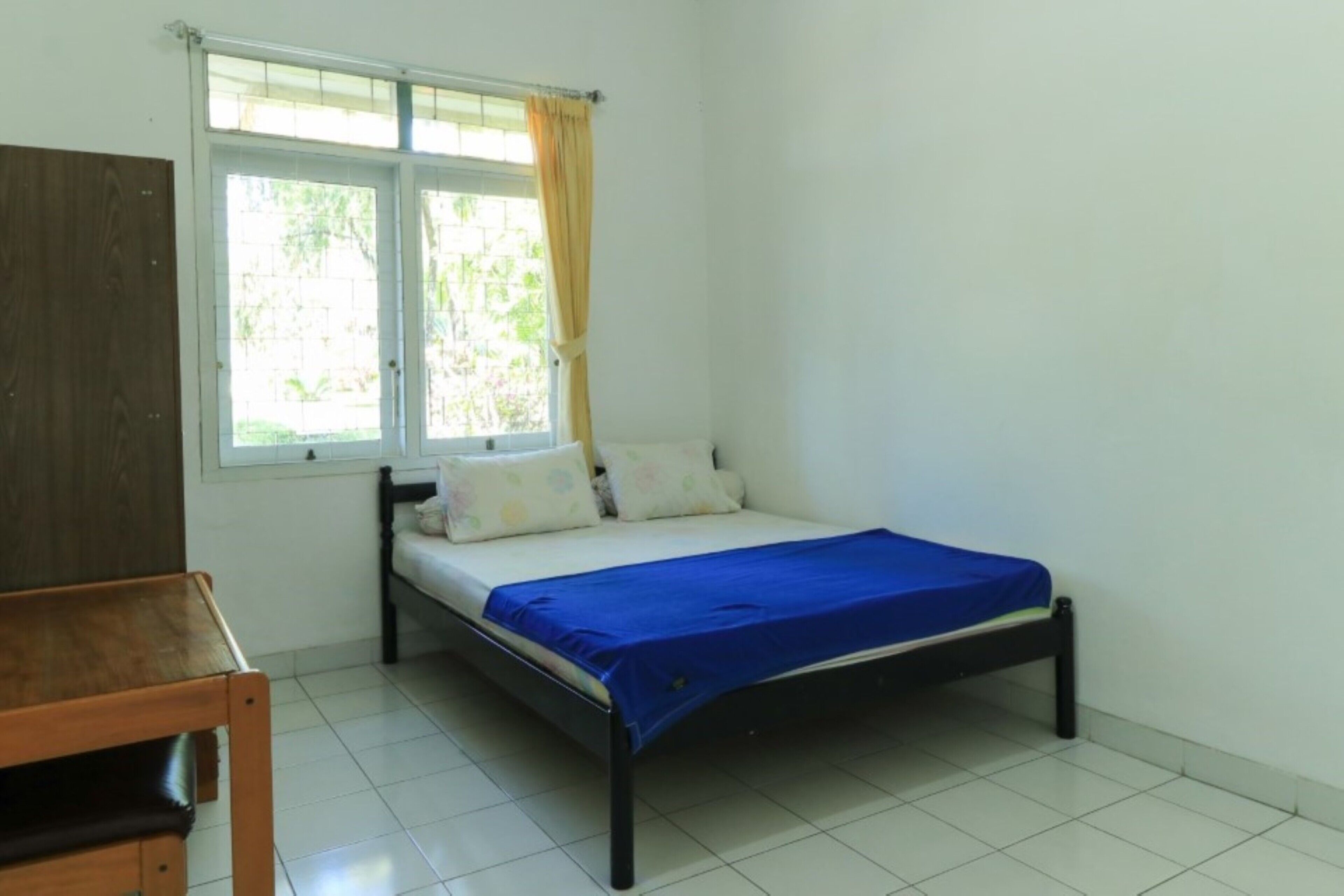 A bedroom with a bed, a window with curtains, a wooden desk, and a tiled floor.