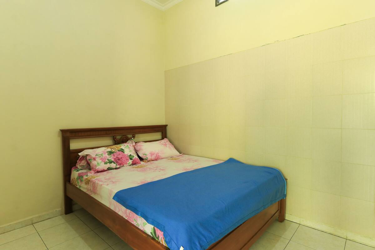 A single bed with a wooden frame and a floral bedspread, positioned against a plain wall with a tiled floor.