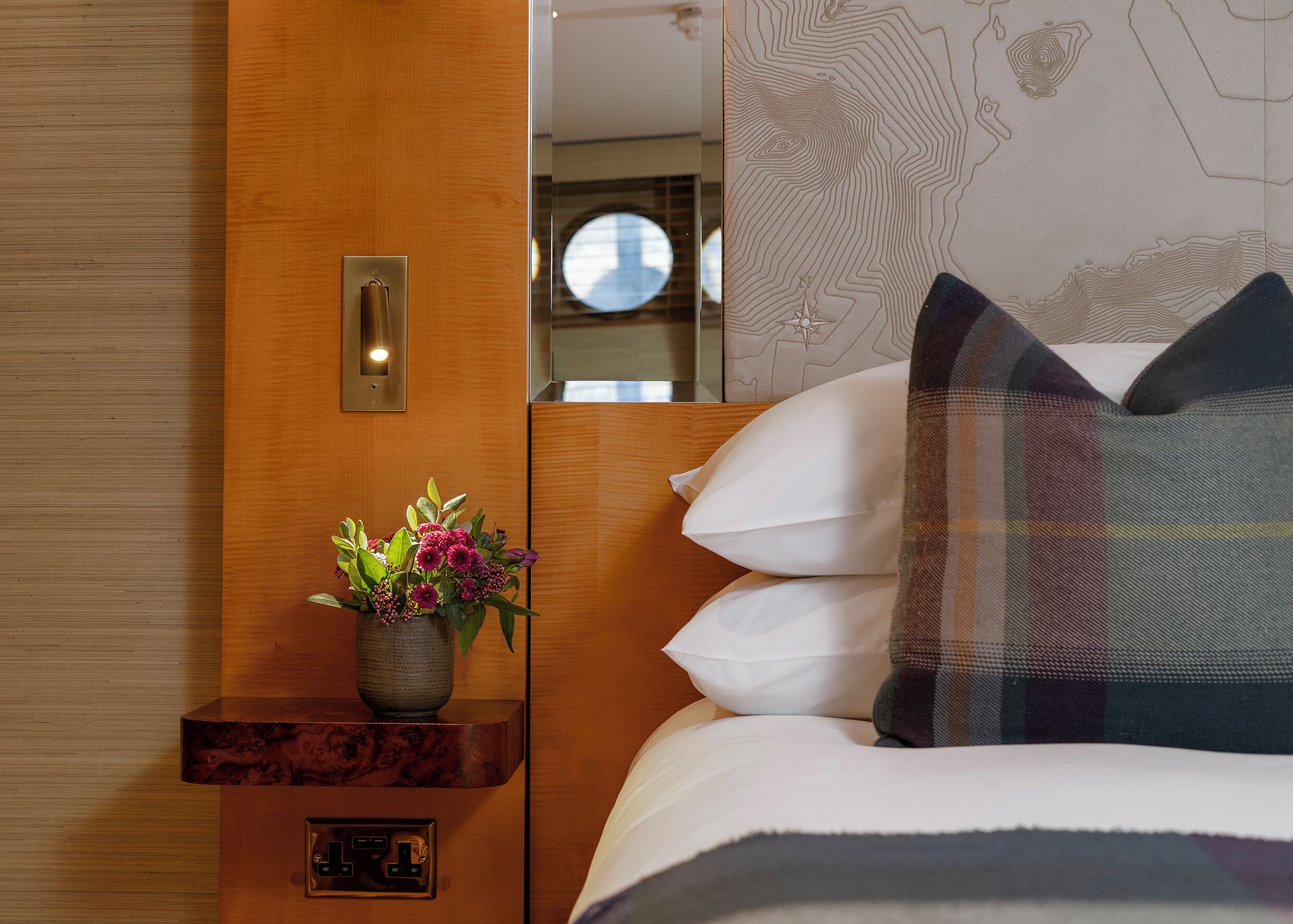 A hotel room with a wooden nightstand, a vase with flowers, a bed with white pillows, and a wall with a map design.