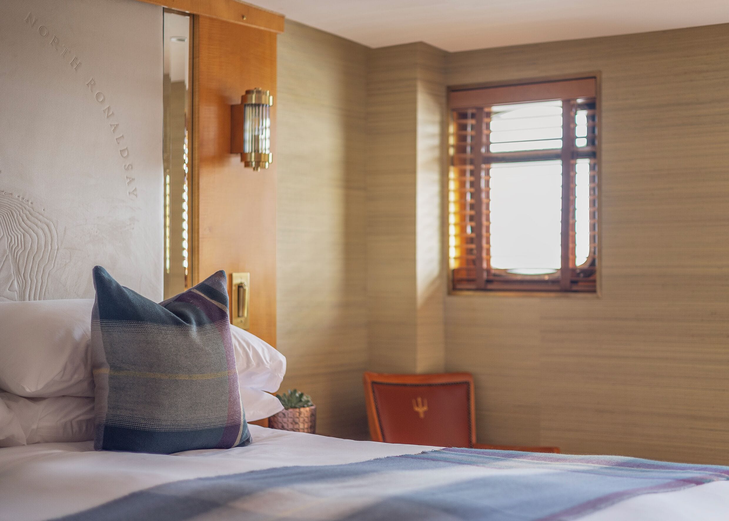 A hotel room with a bed, pillows, a small table with a plant, a chair, and a window with blinds.
