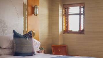 Habitación de hotel con cama, almohadas, una mesita con una planta, una silla y una ventana con persianas.