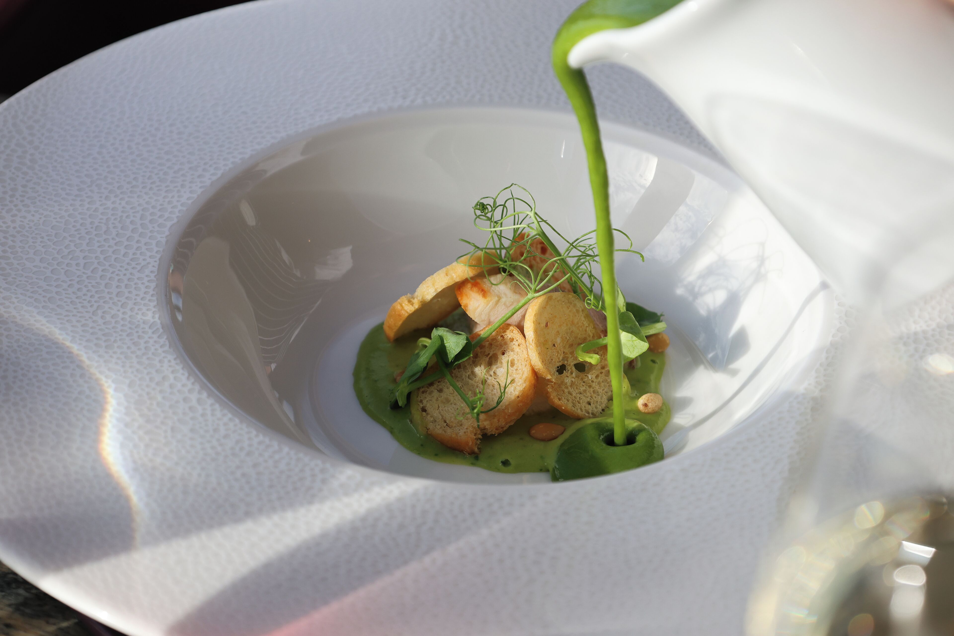 A white plate with a green sauce being poured over a dish with bread and greens.
