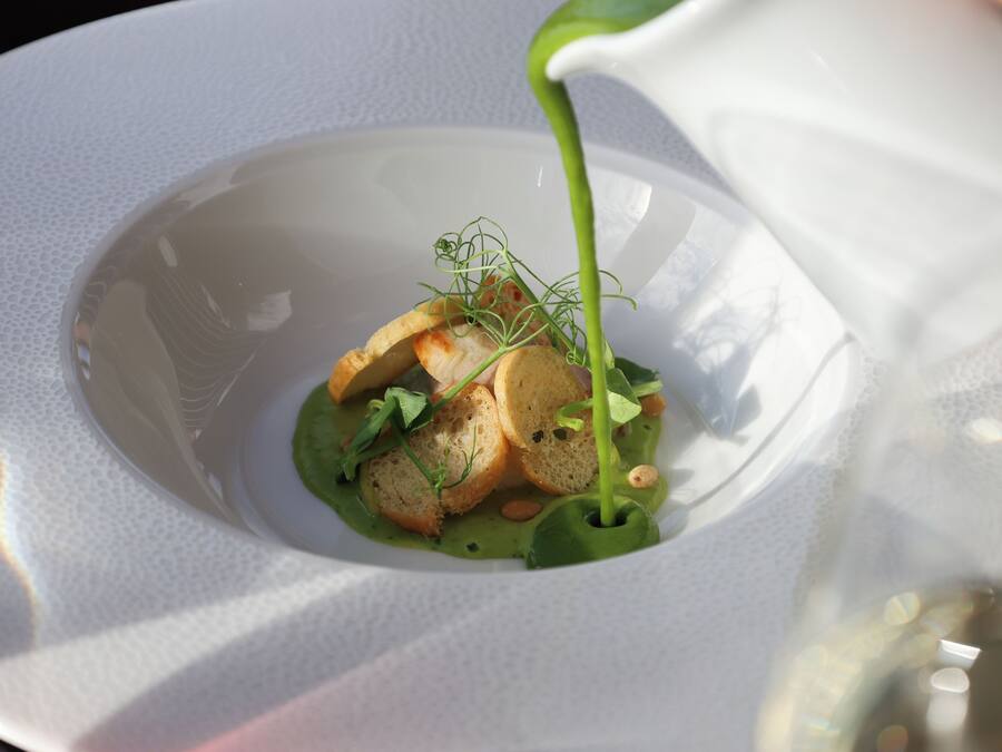 A white plate with a green sauce being poured over a dish with bread and greens.