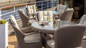 Una terraza con mobiliario de mimbre, una mesa de vidrio y vistas al agua y edificios.