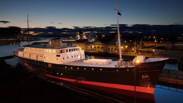 Un barco grande e iluminado amarrado en un puerto, con un edificio al fondo.
