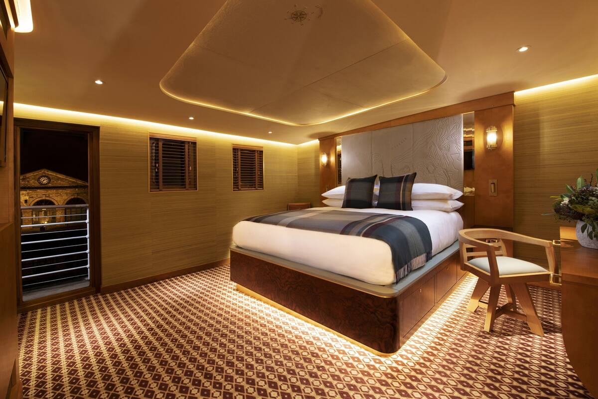 Quarto de hotel moderno com uma cama grande, cabeceira de madeira, piso estampado e uma pequena mesa de escritório com cadeira.
