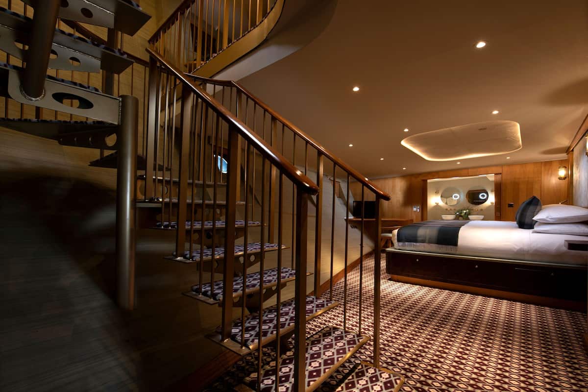 Quarto de hotel moderno com escada em espiral, uma cama grande e um tapete estampado.