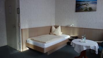 Ein Einzelbett mit Holz-Kopfteil, ein kleiner Nachttisch mit weißem Tischläufer und ein Gemälde mit Meeresblick an der Wand.
