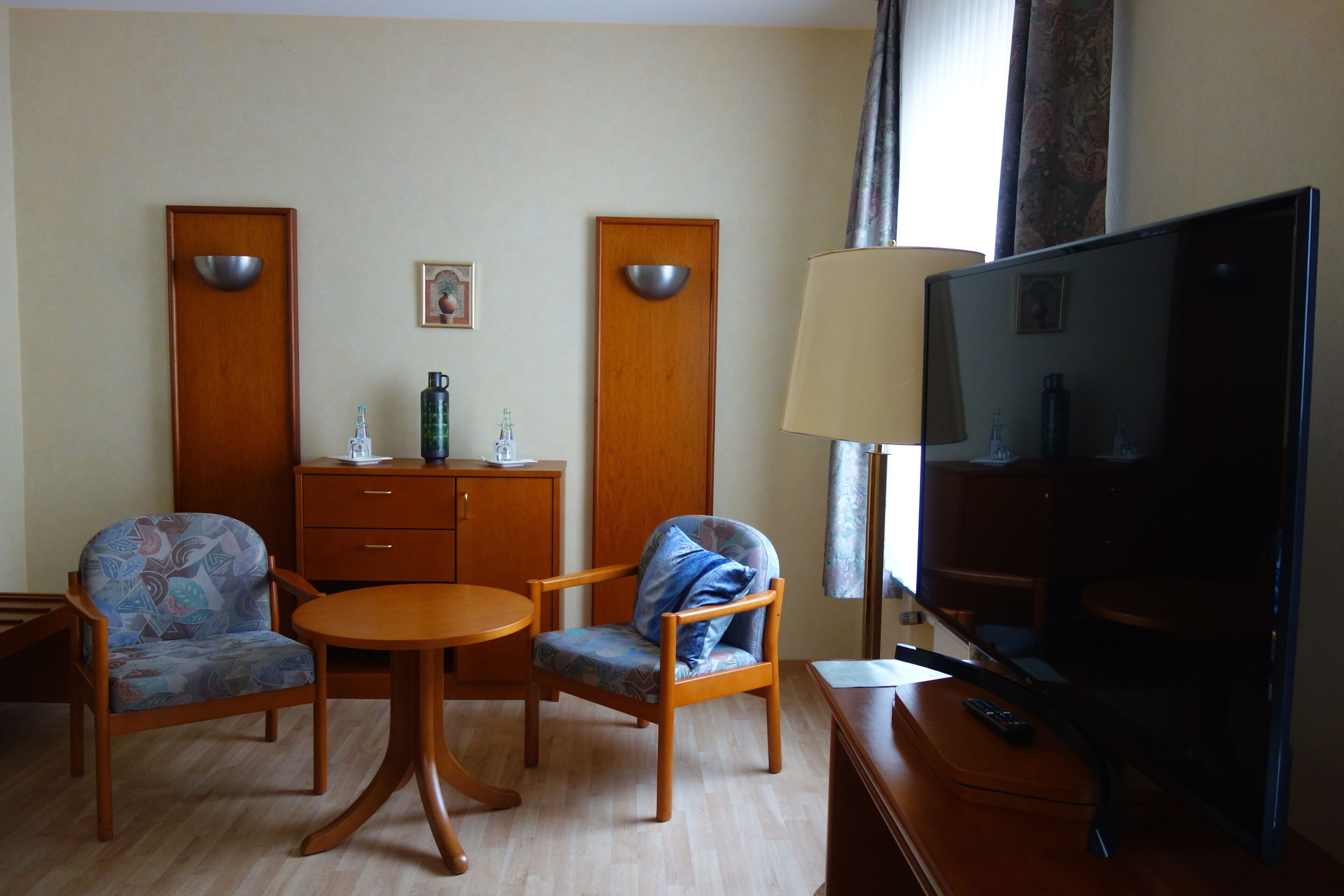 Quarto de hotel com televisão, duas poltronas, uma mesinha, um armário e uma luminária.