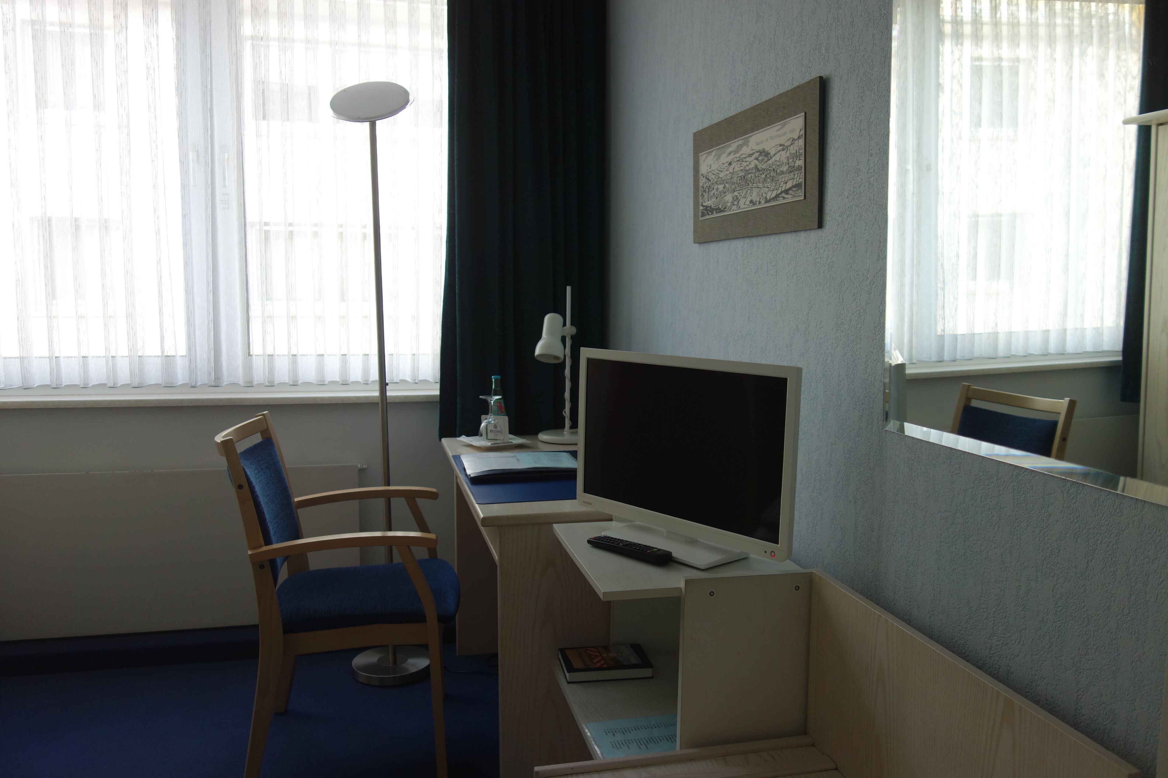 A hotel room with a desk, chair, computer, and a window with sheer curtains.