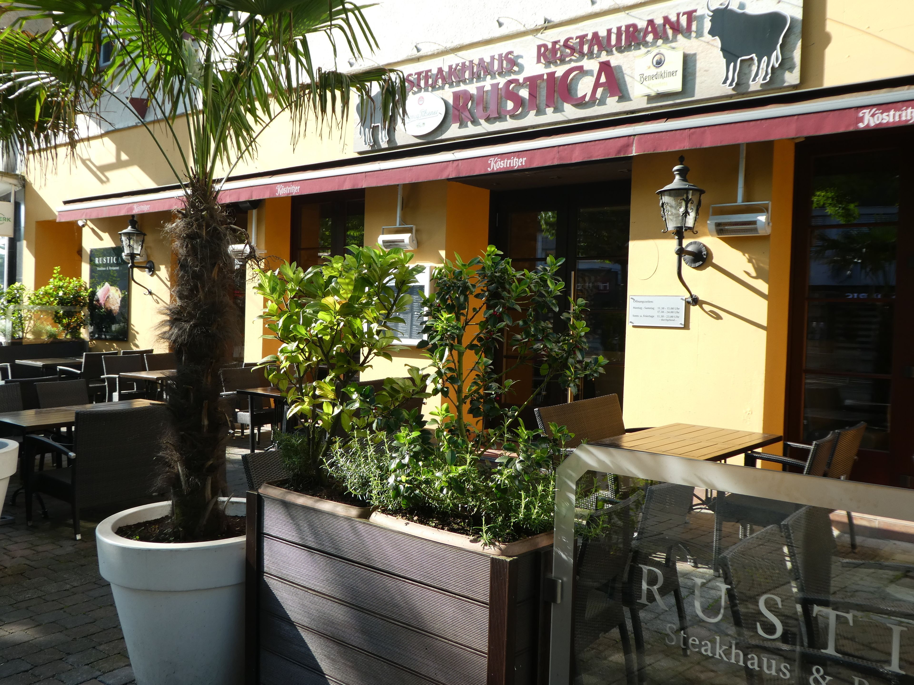 Un área de estar al aire libre con mesas y sillas, una jardinera grande con diversas plantas, y un letrero de restaurante que dice 