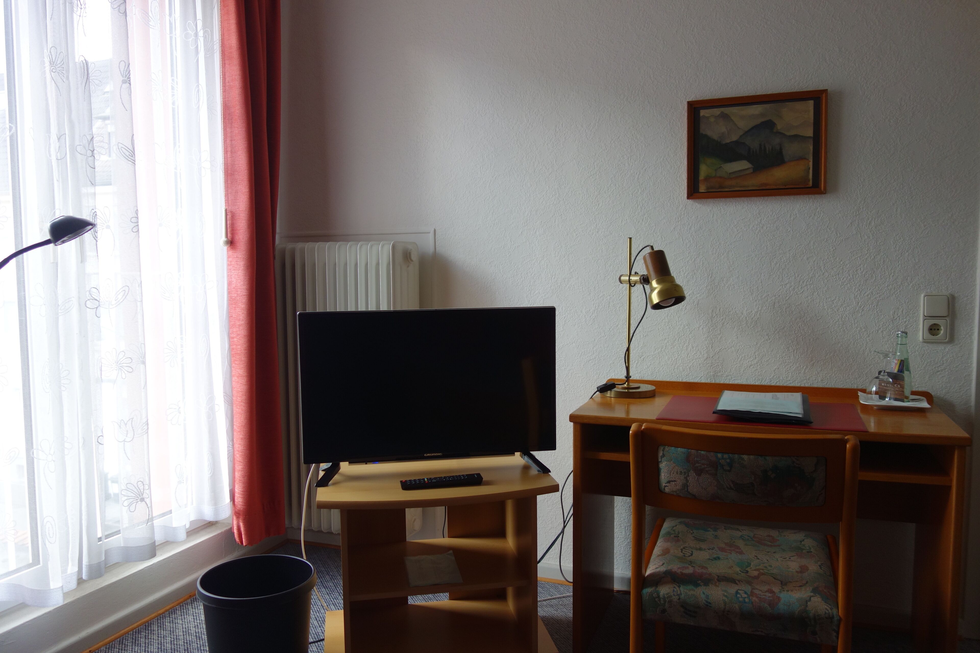 A hotel room with a television on a wooden stand, a desk with a chair, a lamp, and a painting on the wall.