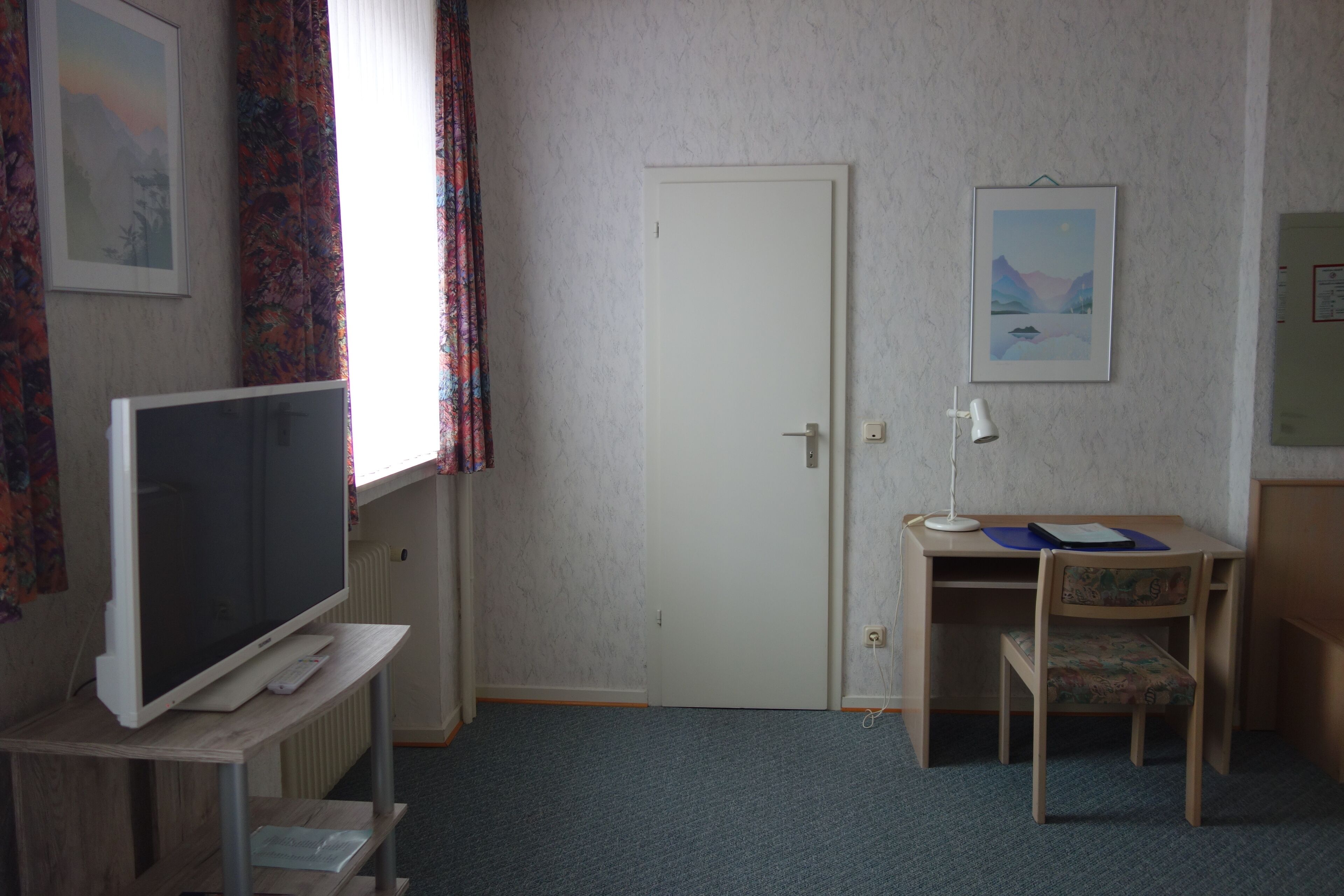 Una habitación con una televisión sobre un mueble bajo, un escritorio con una silla, una puerta y un cuadro enmarcado en la pared.