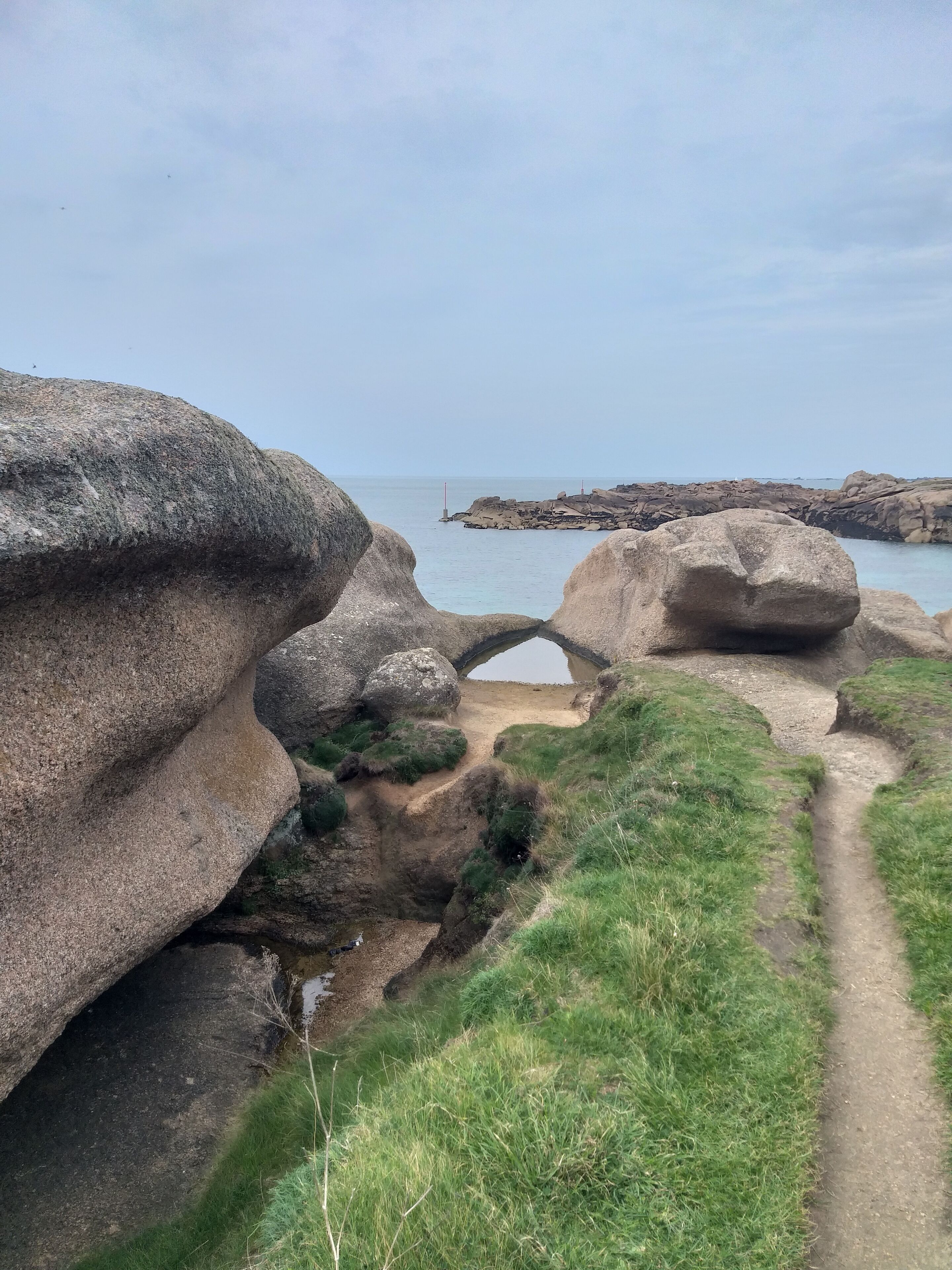 A coastal path with large boulders and a narrow passageway leading to the sea.