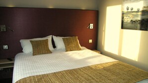 Habitación de hotel con una cama grande, cabecera, mesita de noche y un interruptor de luz en la pared.