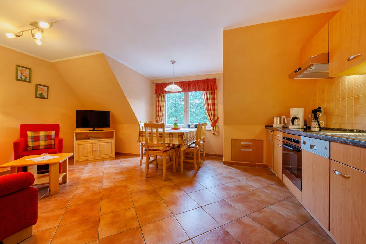 A kitchen with a dining table, chairs, and a TV. There is a window with curtains and a view of trees outside.