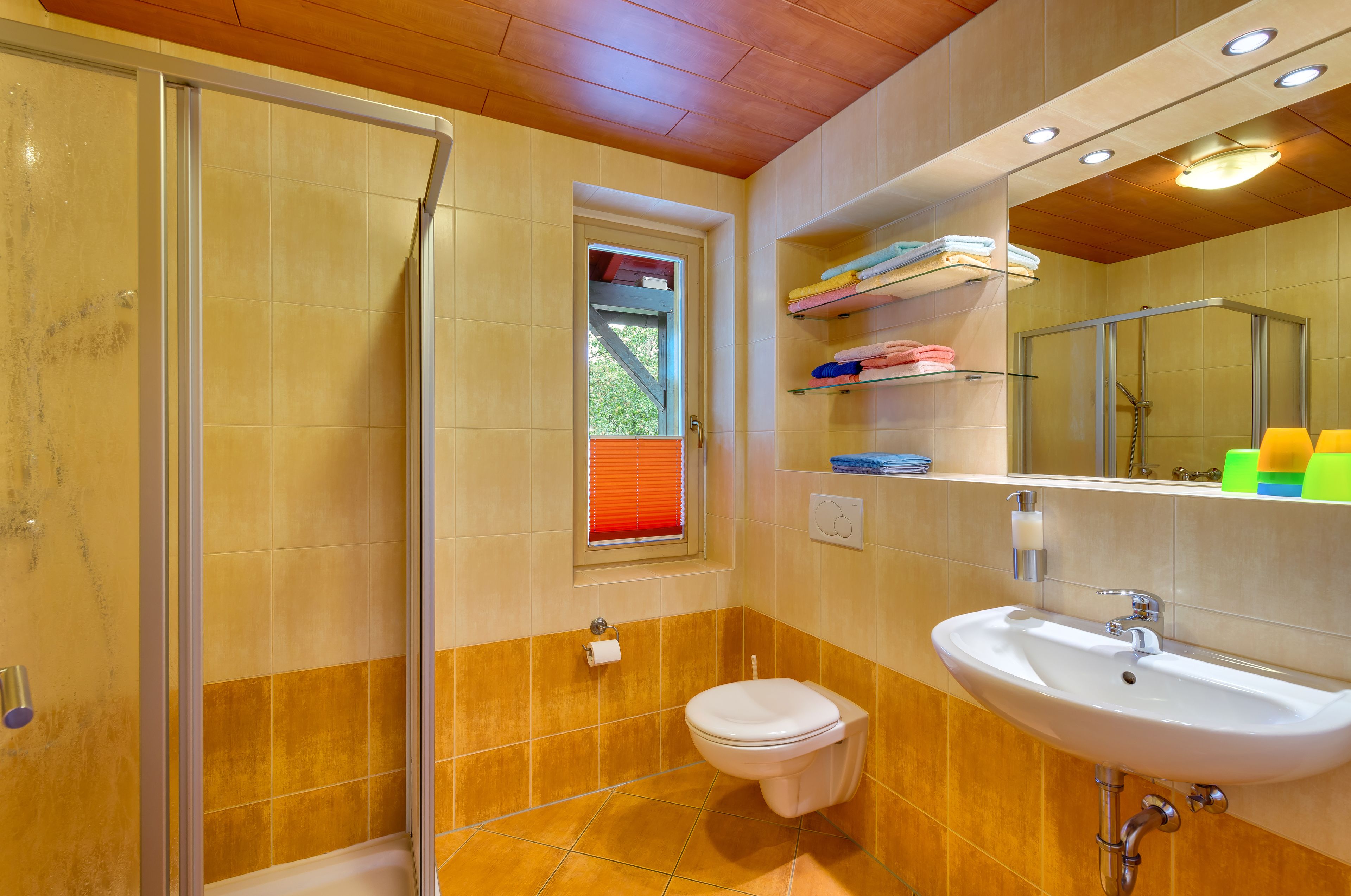 A bathroom with a glass shower enclosure, a toilet, a sink, and a window with blinds.