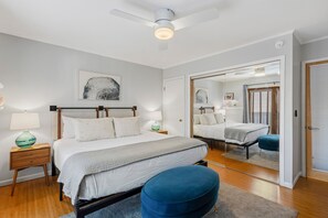 Une chambre moderne avec un lit, des tables de chevet, une commode, un pouf bleu, un ventilateur de plafond et un miroir qui reflète la pièce.