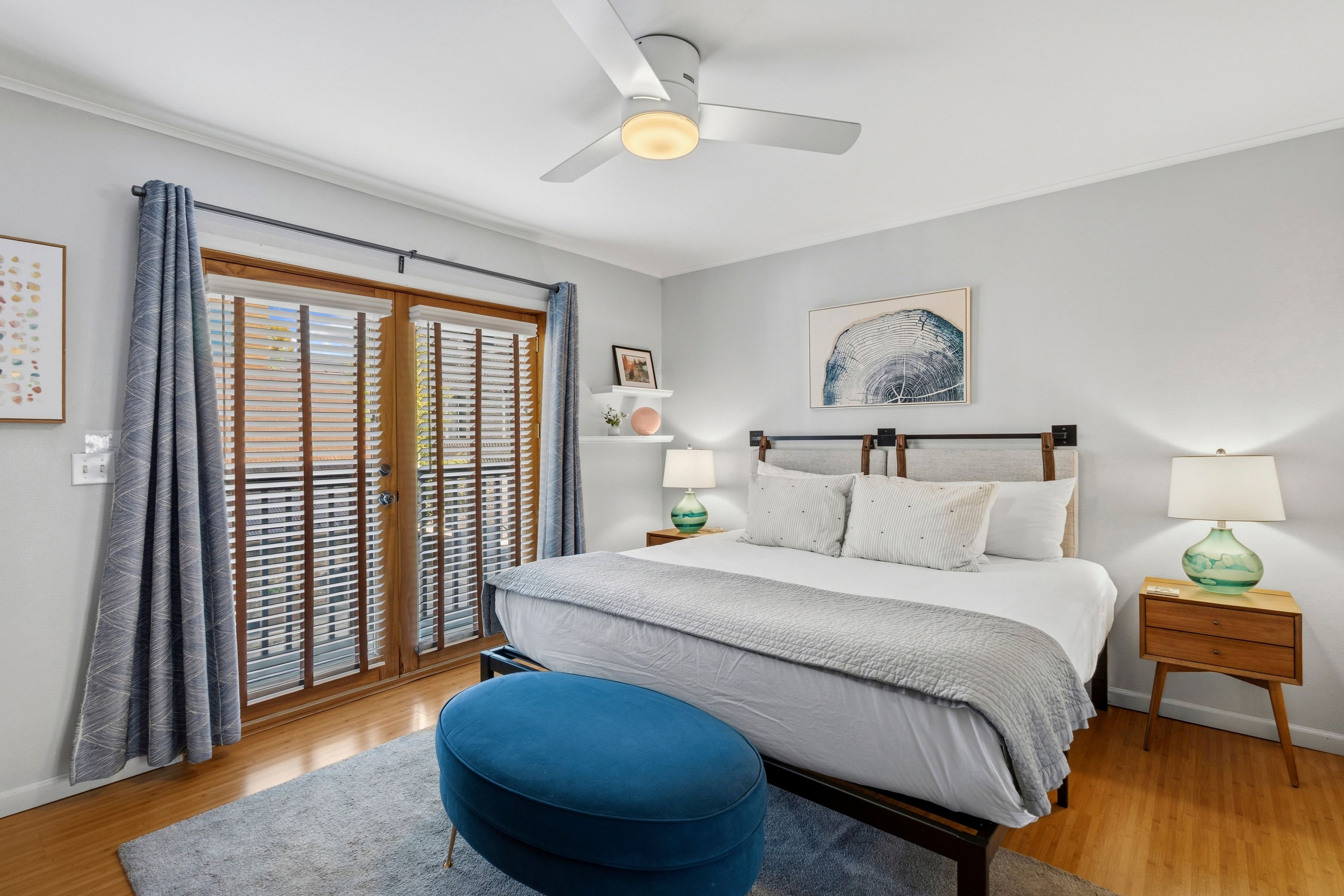 Un dormitorio con cama, mesita de noche, lámpara, ventilador de techo y ventana con persianas.