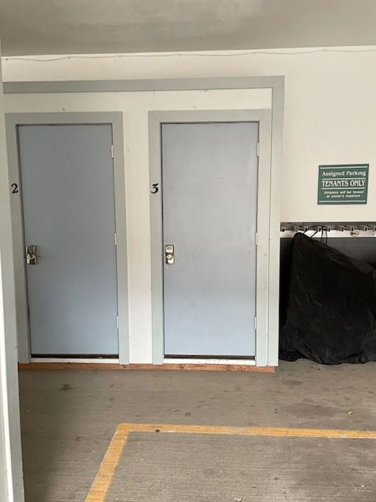 Dos puertas grises con los números 2 y 3, un letrero que indica estacionamiento exclusivo para inquilinos y un objeto negro en el suelo.