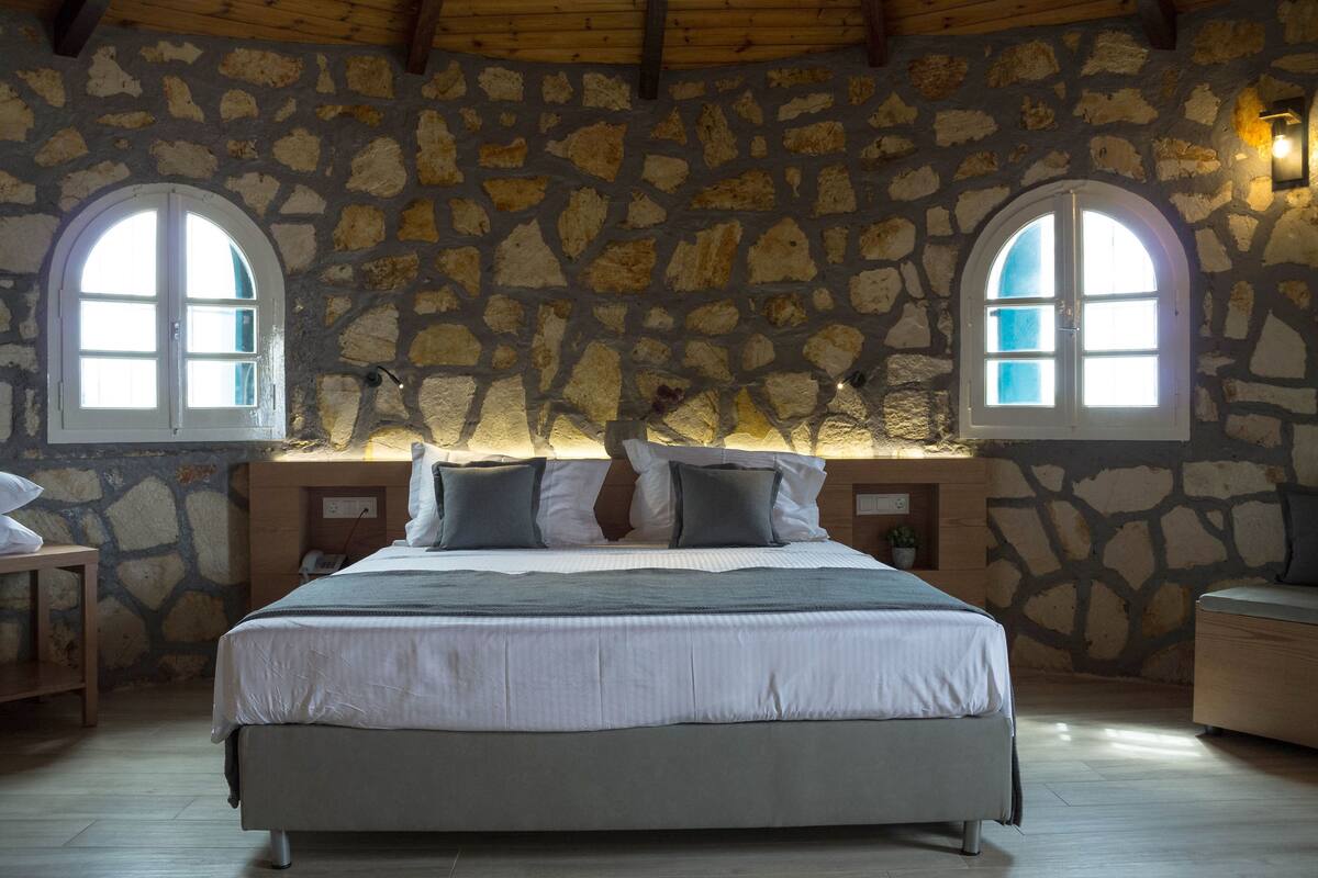 A bedroom with a stone wall, two arched windows, a bed with pillows, and a bedside table with a lamp.