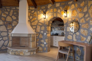 Cozinha com paredes de pedra, lareira embutida, vigas de madeira no teto e área de jantar com mesa e cadeira de madeira.