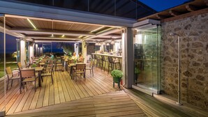 Eine Terrasse im Freien mit Holzboden, Tischen und Stühlen, einer Steinmauer und einer Glaseinfassung.