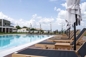 Zona de piscina con sillas de descanso y sombrillas, cielo despejado y edificios al fondo.