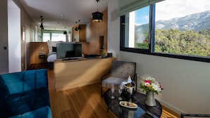 A modern hotel room with a sofa, a dining table with a vase of flowers, a TV, and a view of mountains through the window. - Hotel Sainte Victoire (Vauvenargues)