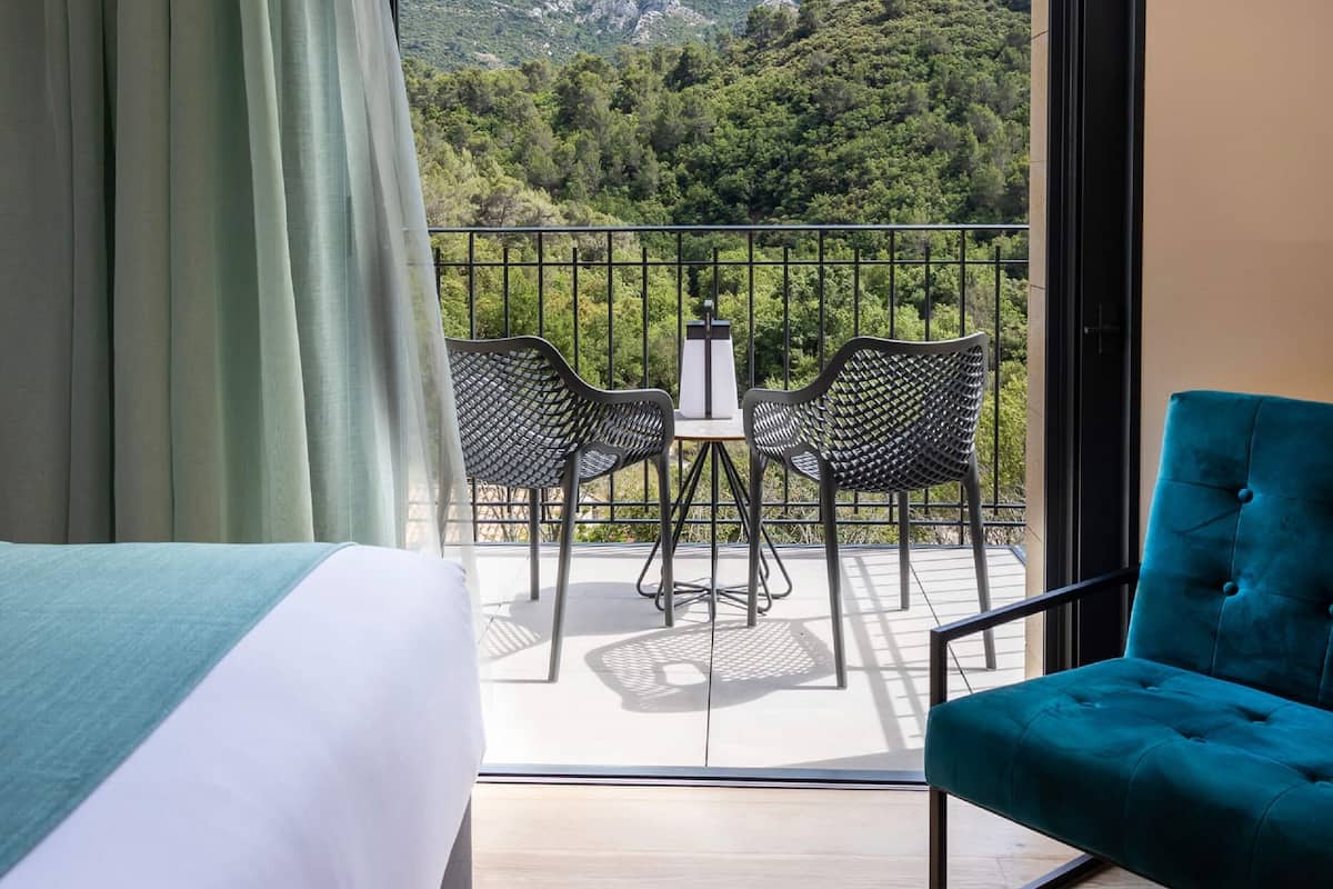 Una habitación de hotel con balcón, una cama, un sillón azul turquesa y una mesa con sillas.