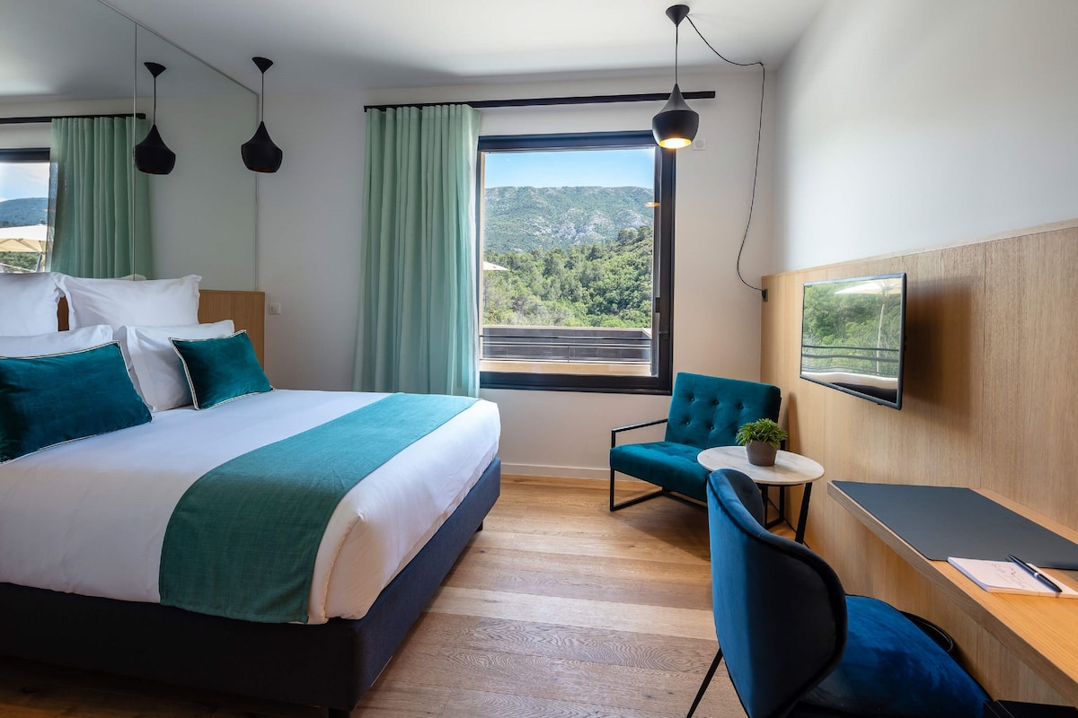 A modern hotel room with a large bed, a desk with a chair, a television, and a view of the mountains.