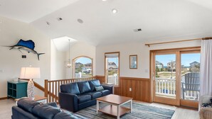 A spacious living room with a large sofa, a coffee table, and a view of the neighborhood through a sliding glass door. - Wise Choice: Semi-oceanfront | Private Pool | Spa (Nags Head)