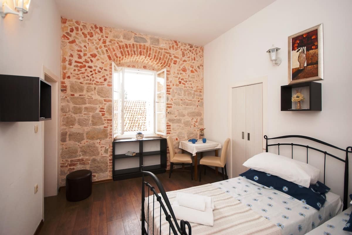 A bedroom with a stone wall, a bed with a floral bedspread, a small table with chairs, and a television on the wall.
