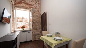 Una pequeña cocina con pared de piedra, una ventana con vista a edificios, una mesa de comedor dispuesta con platos y vasos, y un televisor montado en la pared.