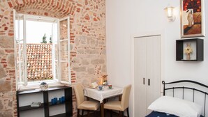 Una habitación pequeña con pared de piedra, ventana con vista a un tejado de tejas, cama, mesa con sillas y estante con libros y tazas.