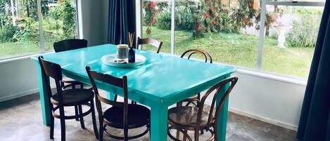 A dining area with a turquoise table and dark wooden chairs, large windows with a view of a garden.