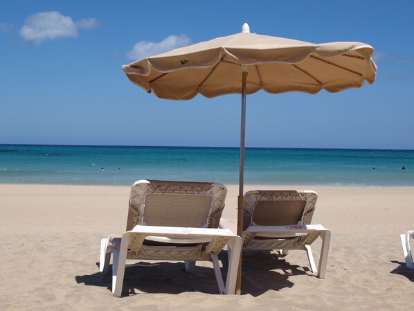 Two beach chairs under a beige umbrella on a sandy beach with a clear blue ocean in the background. - Apartment with balcony and sea view, close to the beach (Costa Calma)