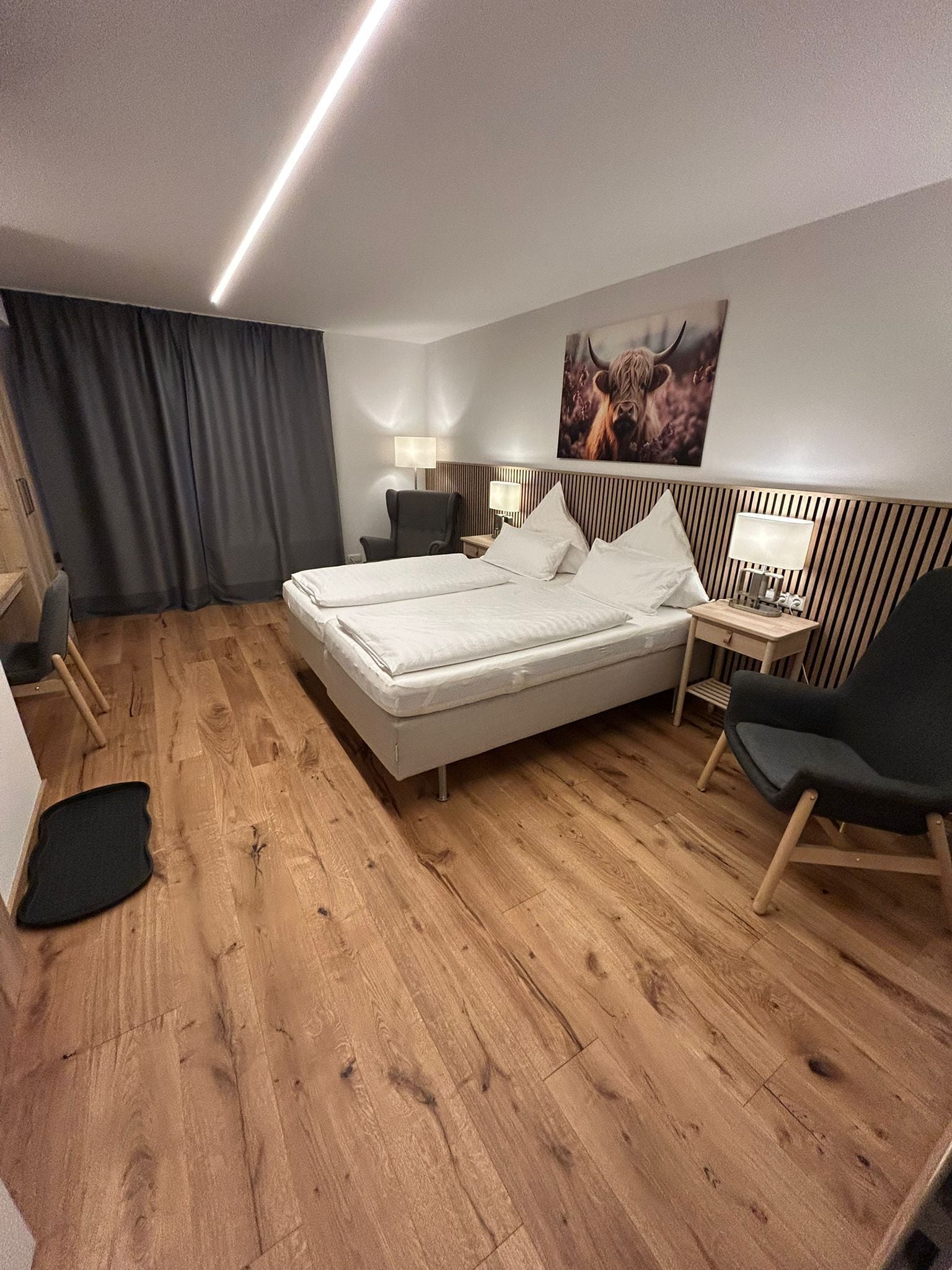 Ein Hotelzimmer mit einem großen Bett, zwei Nachttischen, einem Sessel, einem Schreibtisch, einer Lampe, einem Gemälde und einem Vorhang.
