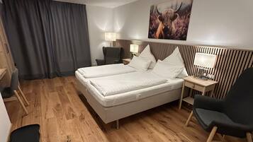 Ein Hotelzimmer mit einem großen Bett, zwei Nachttischen, einem Sessel, einem Schreibtisch, einer Lampe, einem Gemälde und einem Vorhang.