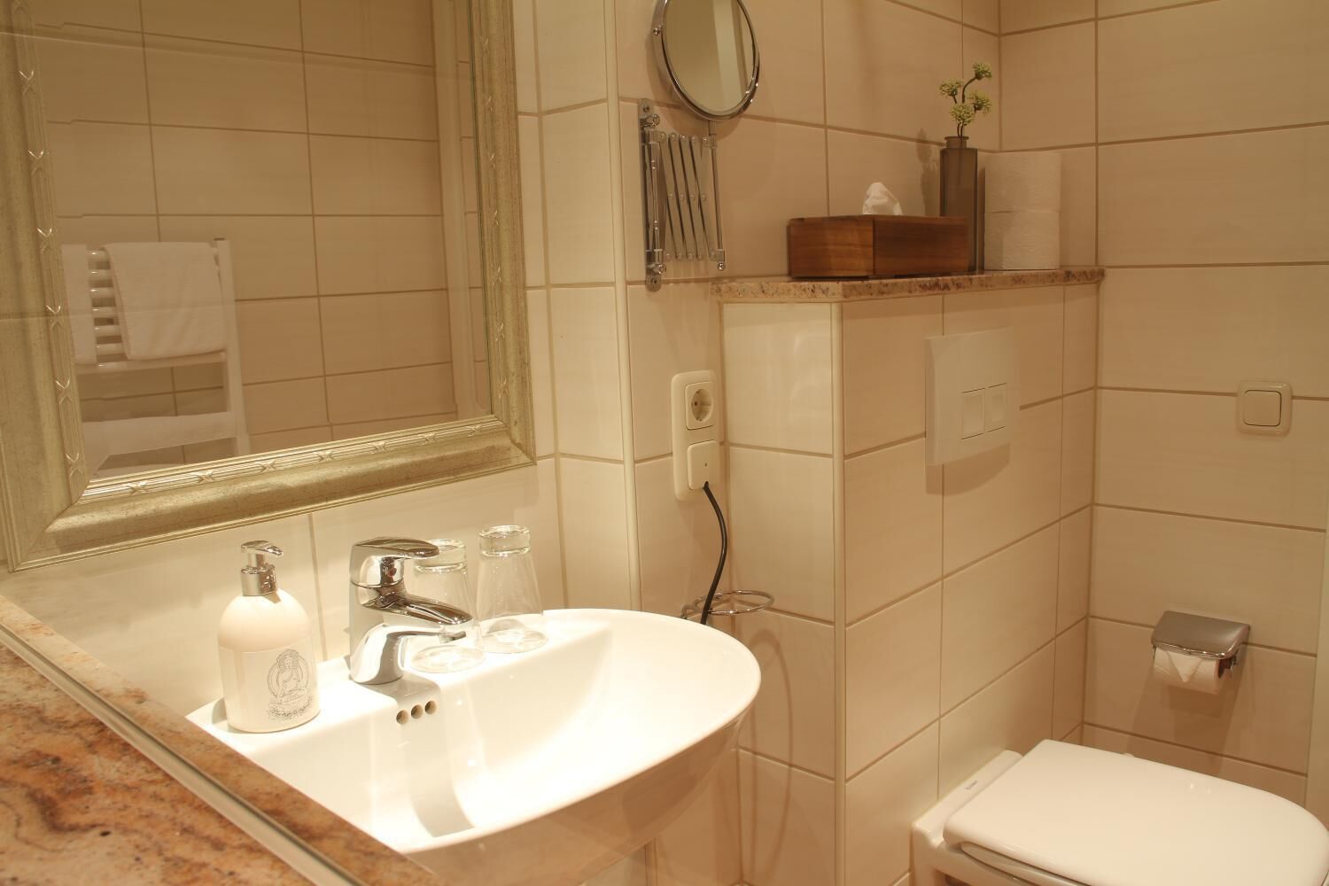 Baño con lavamanos blanco, espejo e inodoro. Se observa un toallero con una toalla y una pequeña estantería con artículos de aseo.
