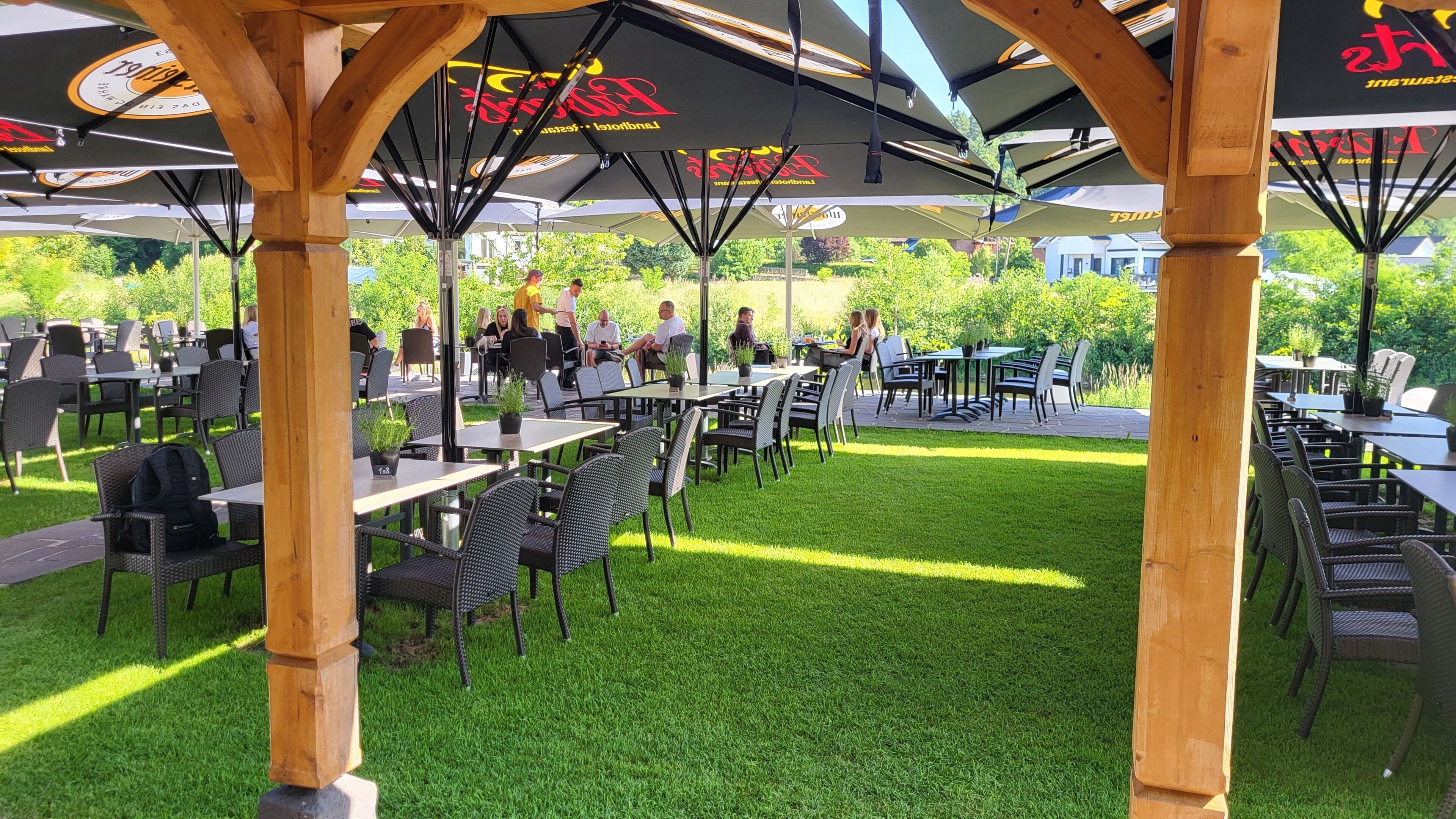 An outdoor patio with tables and chairs, covered by umbrellas, and surrounded by greenery.