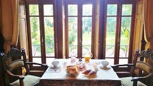 A dining table set with bread, croissants, and a pitcher of orange juice, with a view of a garden through large windows. - CHATEAU DE LA VERNEDE (Saint-Rémy-de-Chargnat)