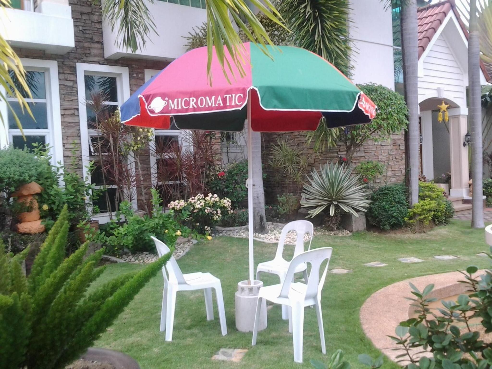 Uma área de estar externa com duas cadeiras de plástico brancas e uma mesa, sob um guarda-sol colorido com a marca 'MICROMATIC'.