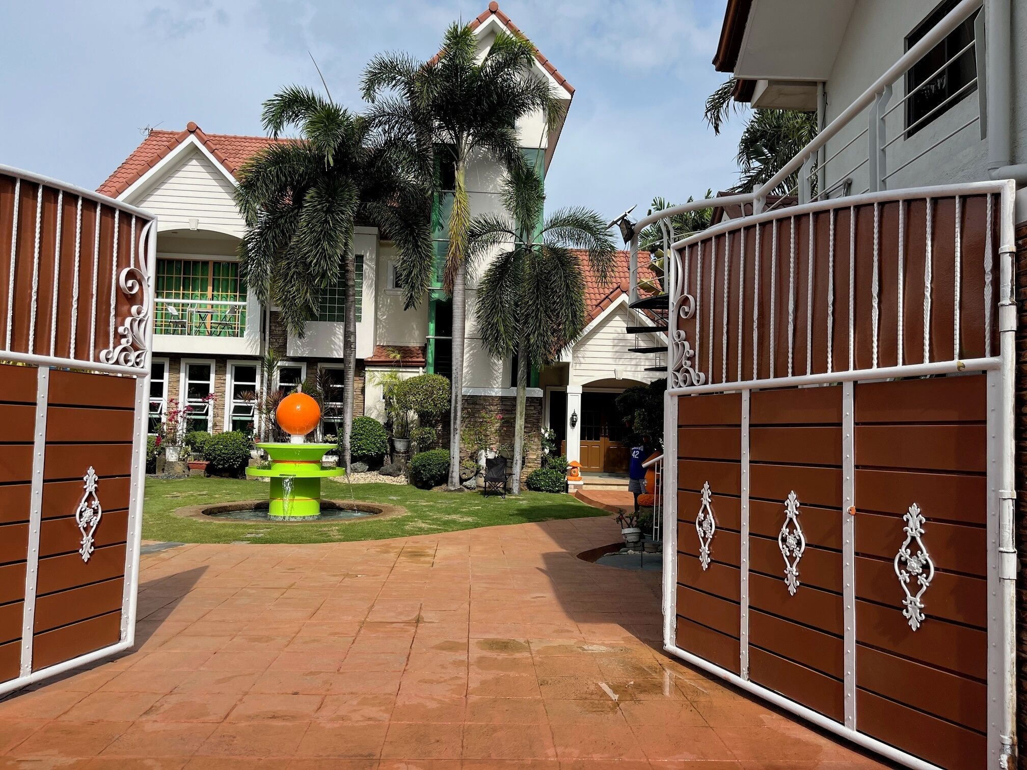 A gated entrance to a property with a fountain, a well-maintained lawn, and a building with a red-tiled roof.