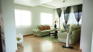 A living room with a green sofa, a coffee table, and a fan. There is a window with curtains and a vase with flowers on the coffee table. - Sir Nico Guesthouse and Resort (Plaridel)