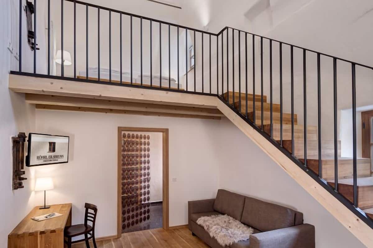 Un loft espacioso con techo alto, una escalera de madera, una zona de estar con sofá y una mesita, y una lámpara fijada a la pared.