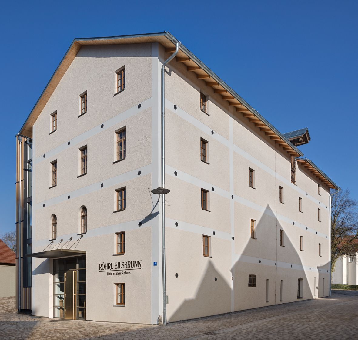 A multi-story hotel building with a beige facade, multiple windows, and a sign indicating it is the "Röhrl EISBRUNN Hotel im Alplhaus."