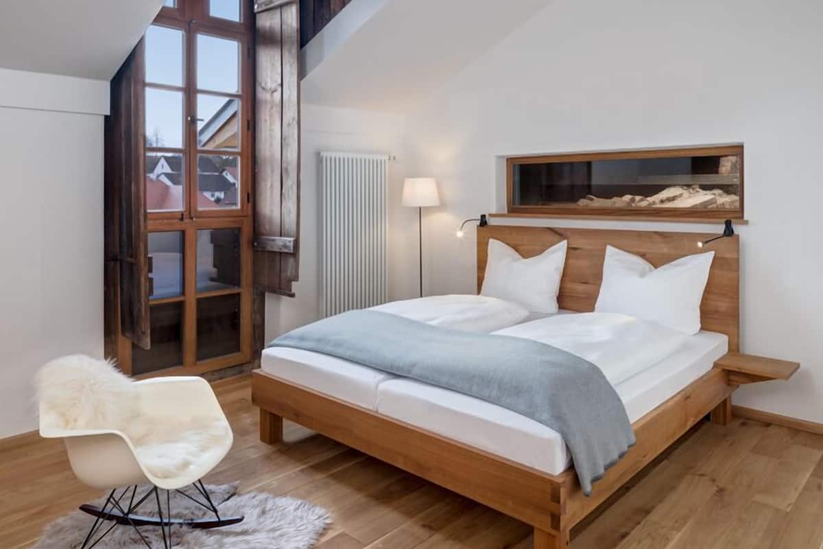 Un dormitorio con una cama de madera, una silla blanca, un manto de piel y una ventana con vistas a edificios.